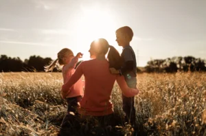 A mom kneeling, hugs her two kids in a open field.