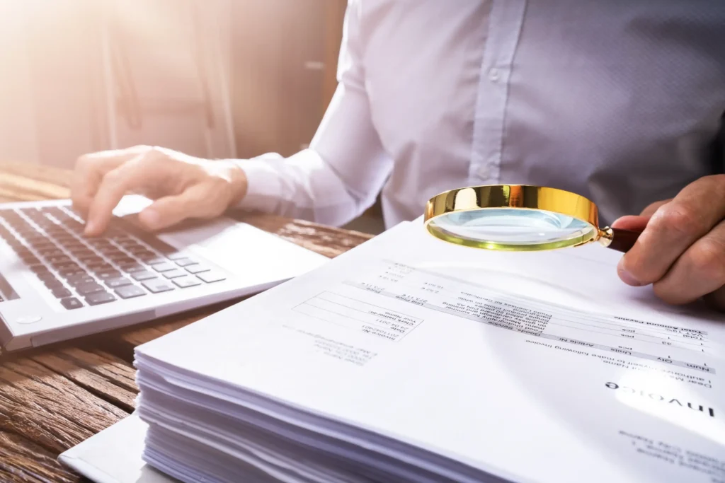 A woman with a magnifying glass looking at a stack of papers and working on a laptop.